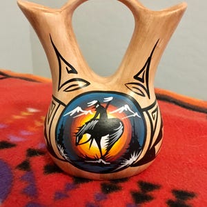 Pode incluir: Um vaso de cerâmica bege com uma alça e dois bicos. O vaso apresenta um desenho pintado de um pássaro em voo contra um pôr do sol, com montanhas e árvores. Padrões geométricos pretos adornam os lados.