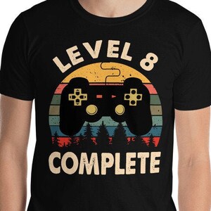 May include: Black t-shirt with a retro-style graphic of a video game controller and the text "Level 8 Complete".