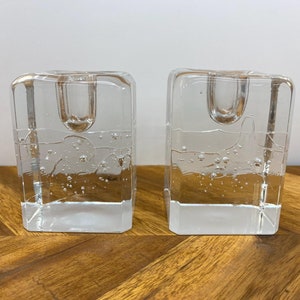 Peut inclure: Deux bougeoirs en verre transparent avec une surface texturée et bullée. Les bougeoirs sont de forme carrée avec une base rectangulaire.