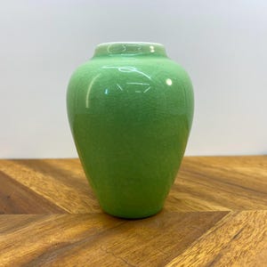 Puede incluir: Un pequeño jarrón de cerámica verde con un esmalte texturizado. El jarrón tiene un cuerpo redondeado y un cuello estrecho.