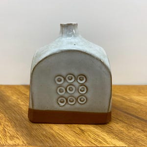 Puede incluir: Un pequeño jarrón de cerámica blanca con un borde marrón y un patrón de nueve círculos en la parte delantera. El jarrón está sobre una superficie de madera.