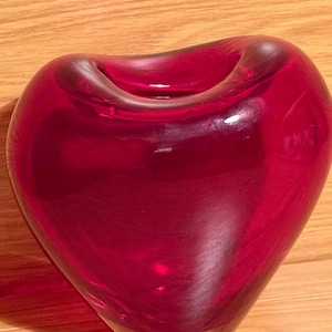 Peut inclure: Un vase en verre rouge rubis en forme de cœur. Le vase a une finition lisse et brillante et une large ouverture en haut. Le vase est posé sur une surface en bois.
