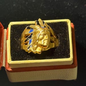 Peut inclure: Une bague dorée représentant un chef amérindien, avec des détails bleus et noirs. La bague est présentée dans un petit coffret ouvert, avec un extérieur rouge et crème et un intérieur noir.