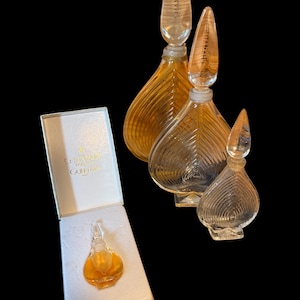 May include: Vintage perfume bottles with clear glass stoppers and amber-colored liquid. The bottles have a leaf-like design and are labeled "CHAMADE". One bottle is in a white box with gold trim and the text "CHAMADE GUERLAIN PARIS".