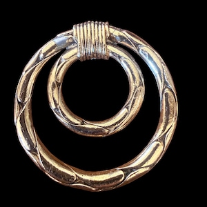 Peut inclure: Broche dorée composée de deux cercles entrelacés. Le plus grand cercle présente un motif texturé, tandis que le plus petit est lisse. Les cercles sont reliés par un élément cylindrique nervuré. Le fond est noir.