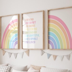 May include: Three framed rainbow prints with positive affirmations, including "I am kind", "I am smart", "I am brave", "I am strong", "I am enough", "I am grateful", "I am capable", and "I am loved". The prints are hung on a white wall above a wooden storage bench with white bins.
