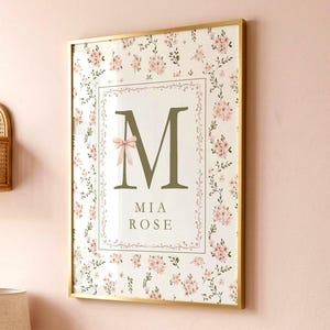 May include: A framed wall print featuring a large olive green letter M with a small pink bow, the name Mia Rose, and a floral pattern of pink flowers and green vines. The print is displayed above a changing pad with a matching floral pattern.