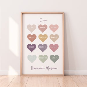 May include: A printable poster with a light brown wooden frame. The poster has a white background with a light pink, peach, and green color scheme. It features a grid of 12 hearts with positive affirmations written in white text. The poster is titled "I am" and signed "Hannah Mason".