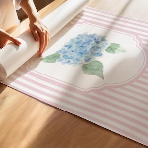 May include: A roll of wallpaper with a floral design featuring a blue hydrangea and green leaves. The wallpaper has a pink and white striped pattern. The image shows the wallpaper being unrolled on a wooden surface.