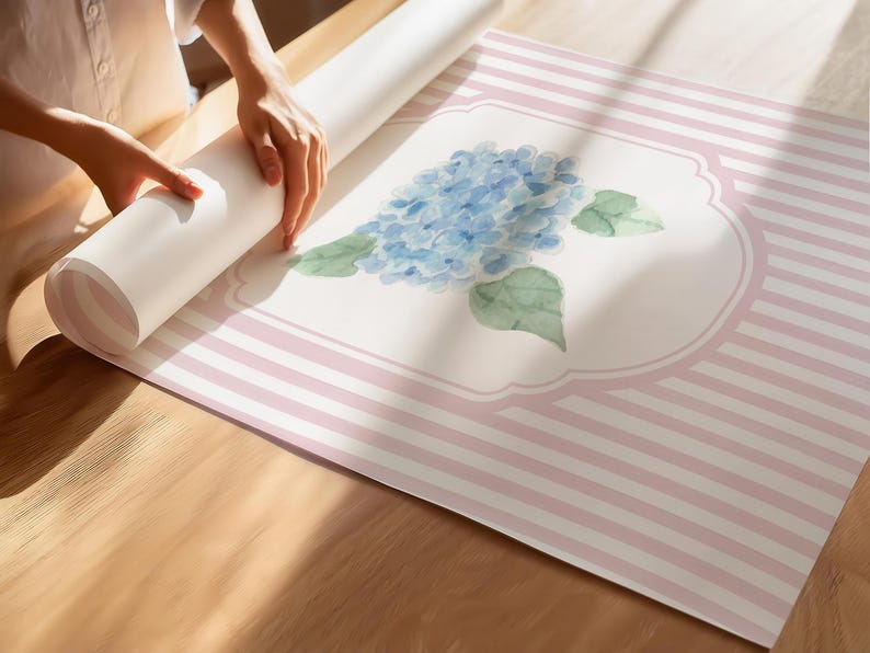 May include: A roll of wallpaper with a floral design featuring a blue hydrangea and green leaves. The wallpaper has a pink and white striped pattern. The image shows the wallpaper being unrolled on a wooden surface.