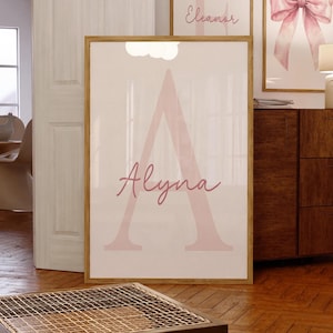 May include: A framed print featuring the name "Alyna" in a soft pink script over a large pink letter "A". The artwork is displayed on a white background, set within a wooden frame. Additional framed art pieces are visible in the background.