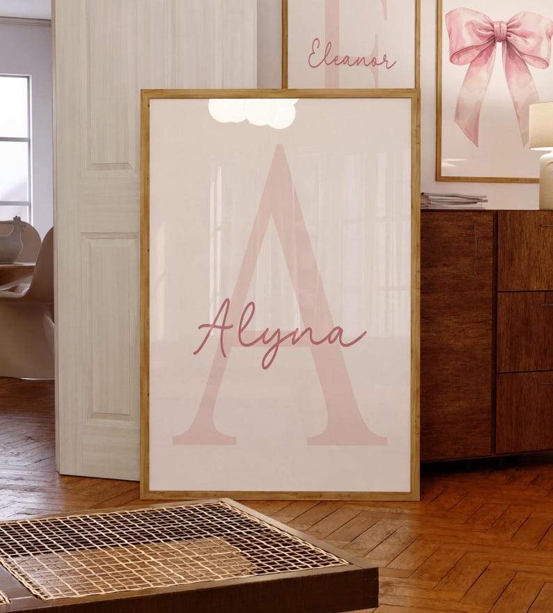 May include: A framed print featuring the name "Alyna" in a soft pink script over a large pink letter "A". The artwork is displayed on a white background, set within a wooden frame. Additional framed art pieces are visible in the background.
