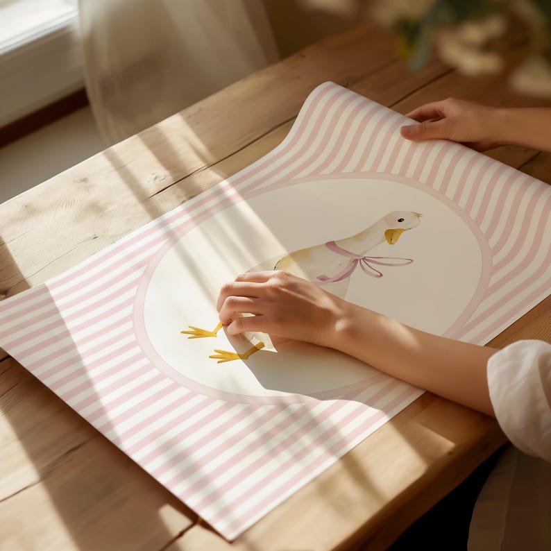 May include: A decorative paper sheet with a watercolor illustration of a goose wearing a pink bow. The goose is centered within a pink oval, surrounded by pink and white stripes. The paper is being held on a wooden surface.
