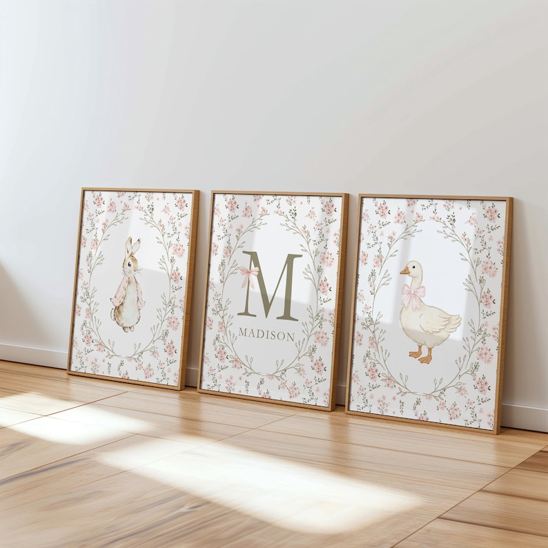 May include: Three framed art prints with a floral wreath design. One features a bunny, another a large letter "M" with the name "Madison", and the third a goose. Each print has a light pink bow and floral accents.