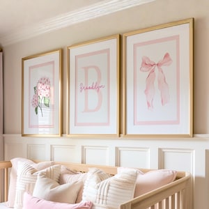 May include: Three framed art prints with gold frames. The prints feature a pink hydrangea, a pink letter "B" with the name "Brooklyn", and a pink bow. The prints are displayed above a crib with pink and white pillows.