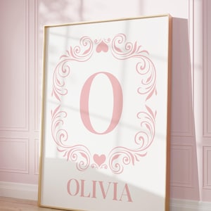 May include: A framed print featuring the name "Olivia" in a soft pink hue. The name is below a large pink letter "O" within a decorative floral and heart-shaped border. The frame is gold-colored.