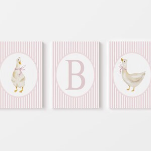 May include: Three framed prints with a nursery theme. Each print features a white circle with a watercolor illustration of a goose wearing a pink bow or a pink letter "B" on a pink and white striped background.