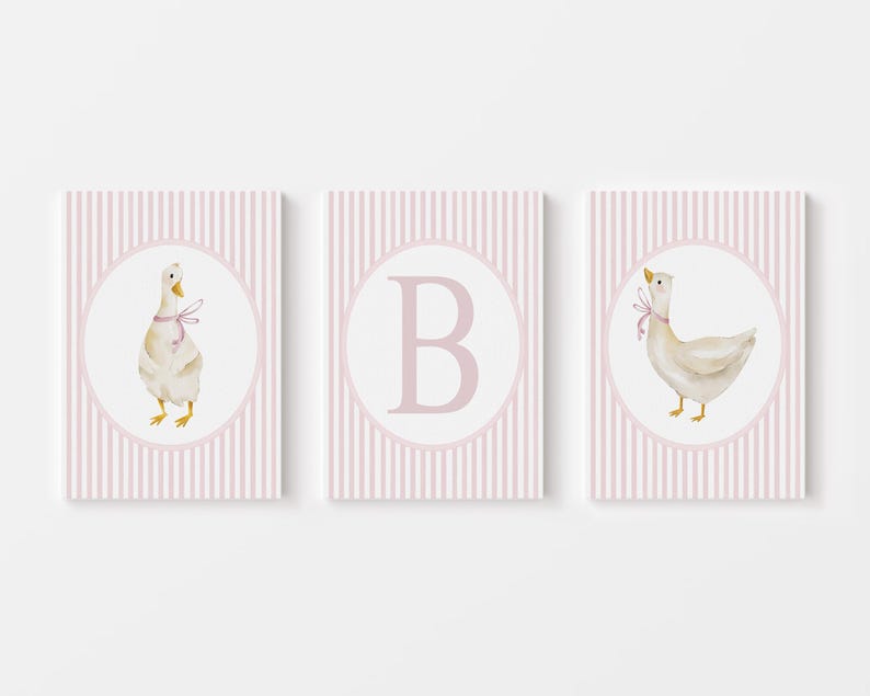 May include: Three framed prints with a nursery theme. Each print features a white circle with a watercolor illustration of a goose wearing a pink bow or a pink letter "B" on a pink and white striped background.
