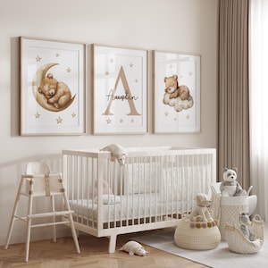 May include: Three framed prints with a nursery theme. The prints feature a brown bear sleeping on a crescent moon, a large letter A with the name "Austin" written below it, and a brown bear sleeping on a cloud. The prints are hung on a white wall above a white crib.