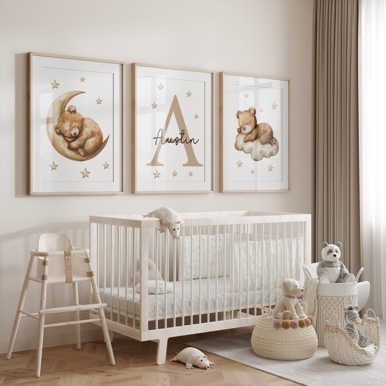 May include: Three framed prints with a nursery theme. The prints feature a brown bear sleeping on a crescent moon, a large letter A with the name "Austin" written below it, and a brown bear sleeping on a cloud. The prints are hung on a white wall above a white crib.