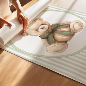 May include: A rolled wallpaper roll featuring a watercolour illustration of a teddy bear with a green bow. The bear is within a white oval with a green border, set against a striped background. The image is on a wooden surface.