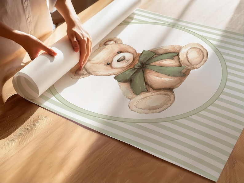 May include: A rolled wallpaper roll featuring a watercolour illustration of a teddy bear with a green bow. The bear is within a white oval with a green border, set against a striped background. The image is on a wooden surface.