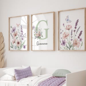 May include: Three framed floral art prints. Each print features watercolor flowers in shades of pink, purple, and lavender, with a butterfly. One print has the letter "G" and the name "Gianna". The frames are light brown.
