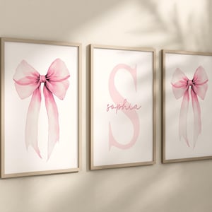 May include: Three framed wall art pieces with a light wood finish. Two feature watercolour paintings of pink bows with long flowing ribbons. The centre frame displays a large pink letter "S" with the name "Sophia" in a script font.