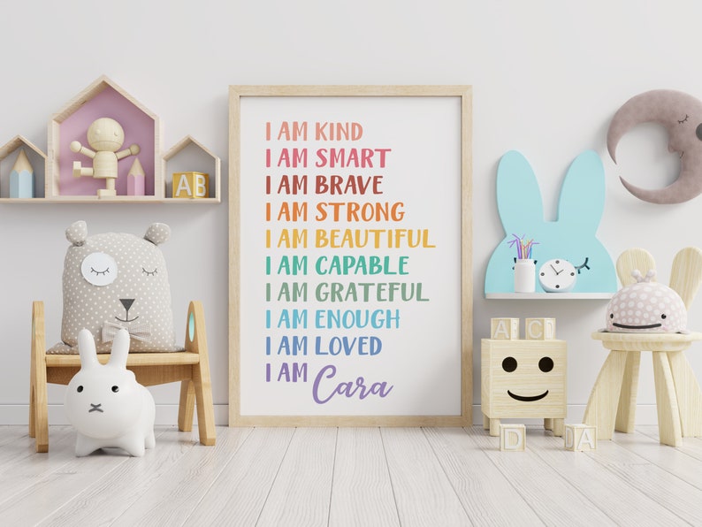 Op de afbeelding: Een kleurrijke poster met de tekst "Ik ben aardig, ik ben slim, ik ben dapper, ik ben sterk, ik ben mooi, ik ben bekwaam, ik ben dankbaar, ik ben genoeg, ik ben geliefd, ik ben Cara" in een regenboogkleurenschema. De poster hangt aan een witte muur in een kinderkamer.