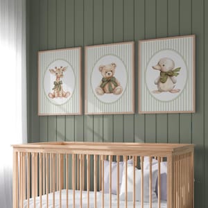 May include: Three framed nursery art prints showing a giraffe, teddy bear, and duckling, each with a green bow. The prints have a striped background and are displayed above a wooden cot. The color palette includes beige, green, and brown.