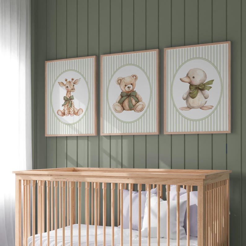 May include: Three framed nursery art prints showing a giraffe, teddy bear, and duckling, each with a green bow. The prints have a striped background and are displayed above a wooden cot. The color palette includes beige, green, and brown.