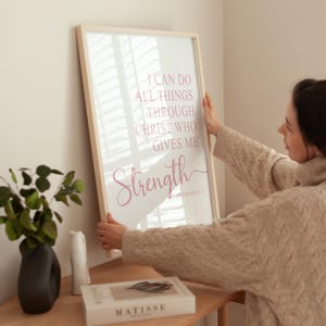 May include: A framed print with the words "I can do all things through Christ who gives me strength" in pink script. The print is held by a person. The frame is light wood.