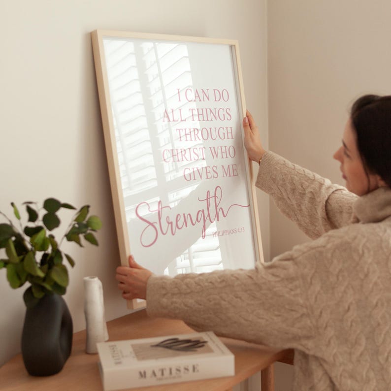 May include: A framed print with the words "I can do all things through Christ who gives me strength" in pink script. The print is held by a person. The frame is light wood.