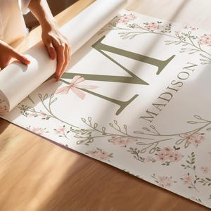 May include: A personalized floral print with the name "Madison" and the letter "M" in olive green. The design includes a floral wreath with pink flowers and a light pink bow. The print is being unrolled on a wooden surface.