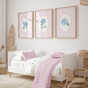May include: Three framed pictures with light wood frames, each displaying a blue hydrangea illustration on a pink striped background. A white bed with pink pillows and a quilted throw is in the foreground, with a wooden rocking horse.