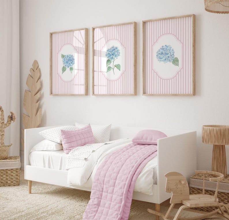 May include: Three framed pictures with light wood frames, each displaying a blue hydrangea illustration on a pink striped background. A white bed with pink pillows and a quilted throw is in the foreground, with a wooden rocking horse.