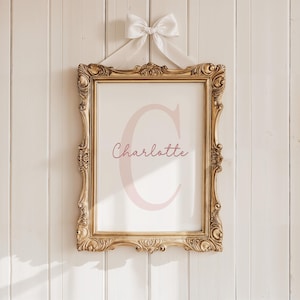 May include: A framed print with the name "Charlotte" in a soft pink script, featuring a large pink letter "C". The ornate gold frame is suspended by a white ribbon tied in a bow, creating a classic and elegant aesthetic.