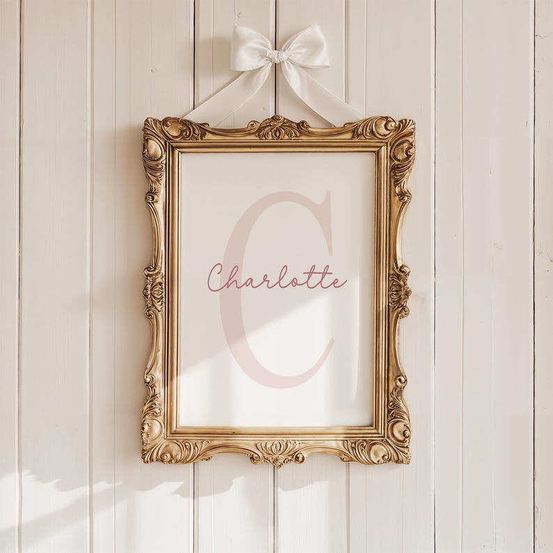 May include: A framed print with the name "Charlotte" in a soft pink script, featuring a large pink letter "C". The ornate gold frame is suspended by a white ribbon tied in a bow, creating a classic and elegant aesthetic.