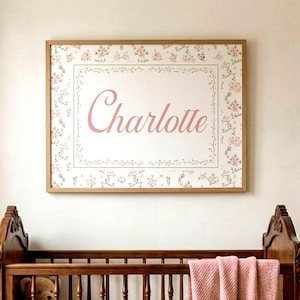 May include: A rectangular wall art print in a light wood frame featuring the name Charlotte in pink script. The print has a white center bordered by a thin line and a surrounding pattern of small pink flowers and green leaves on a white background.