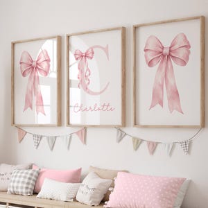 May include: Three framed prints with watercolour pink bows and ribbons. The centre print features the letter "C" and the name "Charlotte". The prints are in light wood frames, with a garland of pennants below.