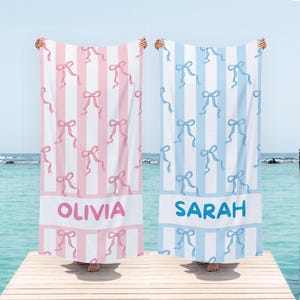 May include: Two beach towels, one pink and one blue, are displayed. The pink towel has vertical stripes and pink bows, with the name "OLIVIA" in pink. The blue towel has blue bows and stripes, with the name "SARAH" in blue.