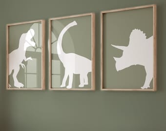 May include: Three framed wall art prints featuring white silhouettes of a Tyrannosaurus Rex, a Brachiosaurus, and a Triceratops against a solid sage green background. Each print is encased in a light-toned wooden frame with a glass front.