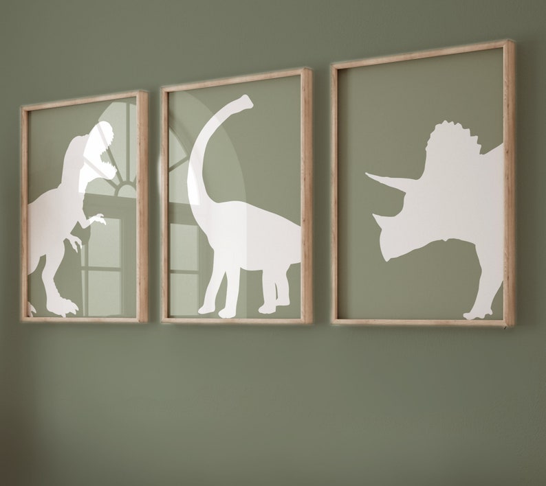 May include: Three framed prints with white silhouettes of dinosaurs on a green wall. The prints are in wooden frames with a light brown finish. The dinosaurs are a triceratops, a stegosaurus, and a tyrannosaurus rex.