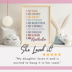 May include: A framed print with the text "I am kind, I am smart, I am brave, I am strong, I am beautiful, I am capable, I am grateful, I am enough, I am loved, I am Nathalie" in colorful text. The print is on a shelf with a white cloud-shaped stuffed toy, a white star-shaped stuffed toy, and a gray and white bunny-shaped stuffed toy.