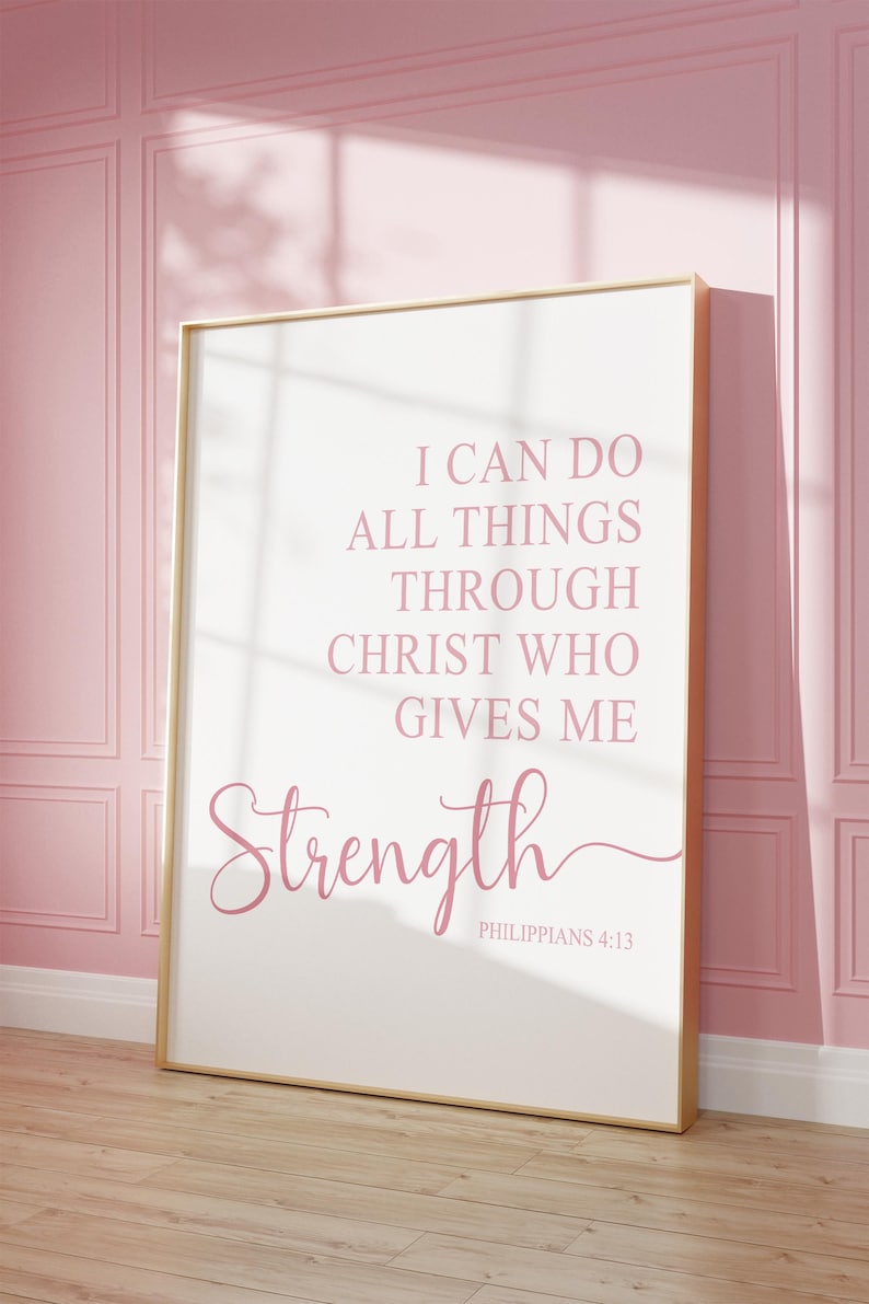 May include: A framed print with a gold border, featuring the text "I CAN DO ALL THINGS THROUGH CHRIST WHO GIVES ME Strength" in pink script. The print is leaning against a pink wall with a wooden floor.