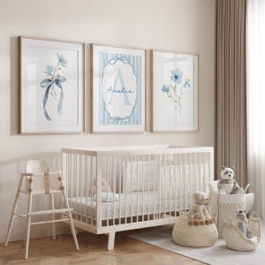 May include: Three framed watercolor prints with light wood frames. The prints feature blue floral designs and the name "Amelia" in script. A white crib, high chair, and baskets with stuffed animals complete the nursery setting.