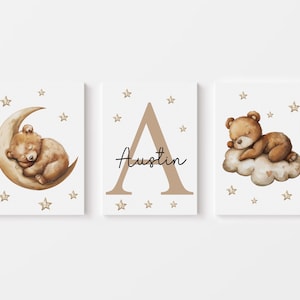 May include: Three nursery prints featuring a brown bear sleeping on a crescent moon, the letter A in brown, and a brown bear sleeping on a cloud. Each print has a white background with gold stars.