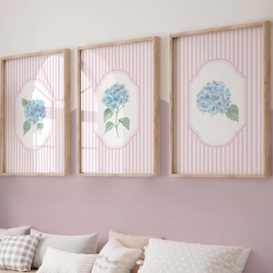 May include: Three framed prints with blue hydrangea flower illustrations. Each print has a light pink striped background and a wooden frame. The hydrangea is set within a white, ornate oval shape.