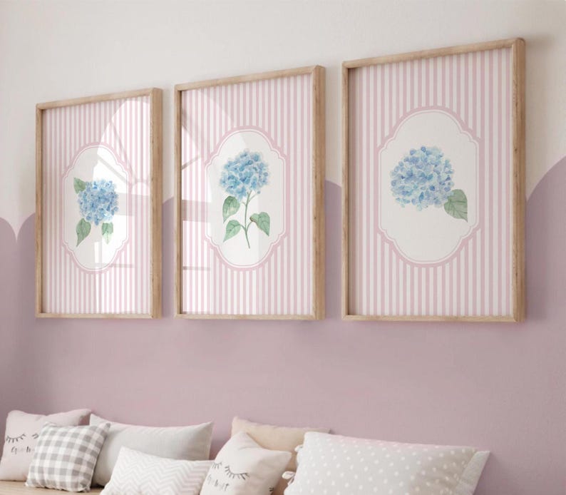May include: Three framed prints with blue hydrangea flower illustrations. Each print has a light pink striped background and a wooden frame. The hydrangea is set within a white, ornate oval shape.