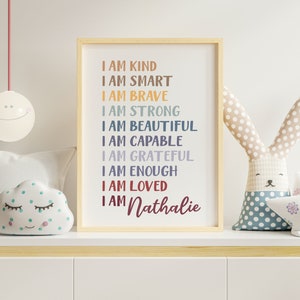 Op de afbeelding: Een ingelijst print met een witte achtergrond en kleurrijke tekst die luidt "Ik ben aardig, ik ben slim, ik ben dapper, ik ben sterk, ik ben mooi, ik ben bekwaam, ik ben dankbaar, ik ben genoeg, ik ben geliefd, ik ben Nathalie". De print staat op een plank met een wit wolkenvormig kussen met groene stippen en een grijs konijnvormig kussen met blauwe stippen.